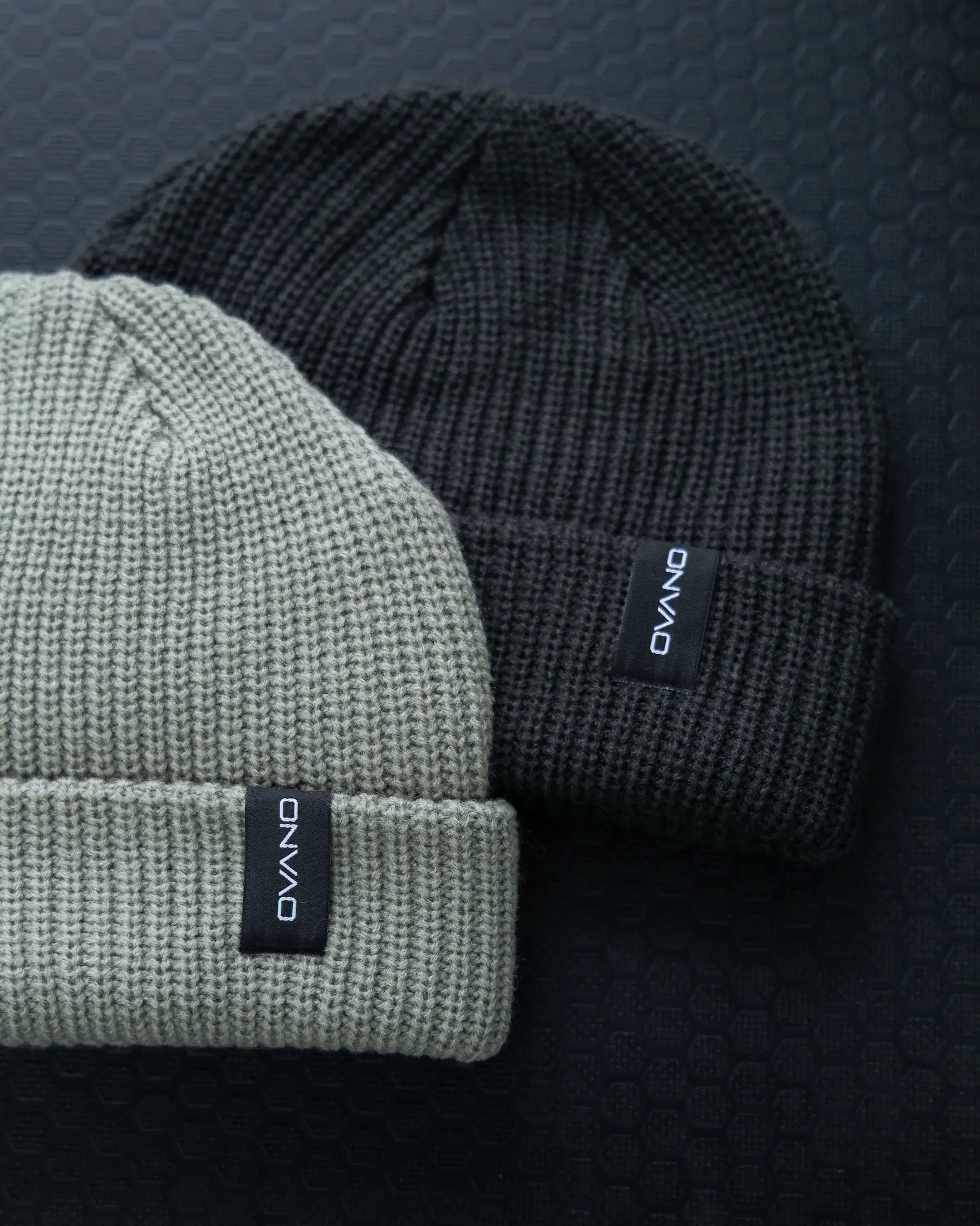Two knit beanie hats, one gray and one black, with a brand label on a textured dark background.