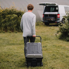 Man pulling a large grey Fridge in a grassy area with a van in the background.