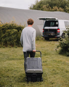 Man pulling a large grey Fridge in a grassy area with a van in the background.