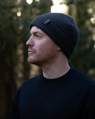 Man wearing a black knit beanie with a Ovano brand logo, standing in a forest setting.